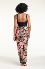 A woman modeling a black one piece swimsuit and a wrap sarong with a vibrant pink, orange, and green floral print, featuring a high side slit and a tied waist.