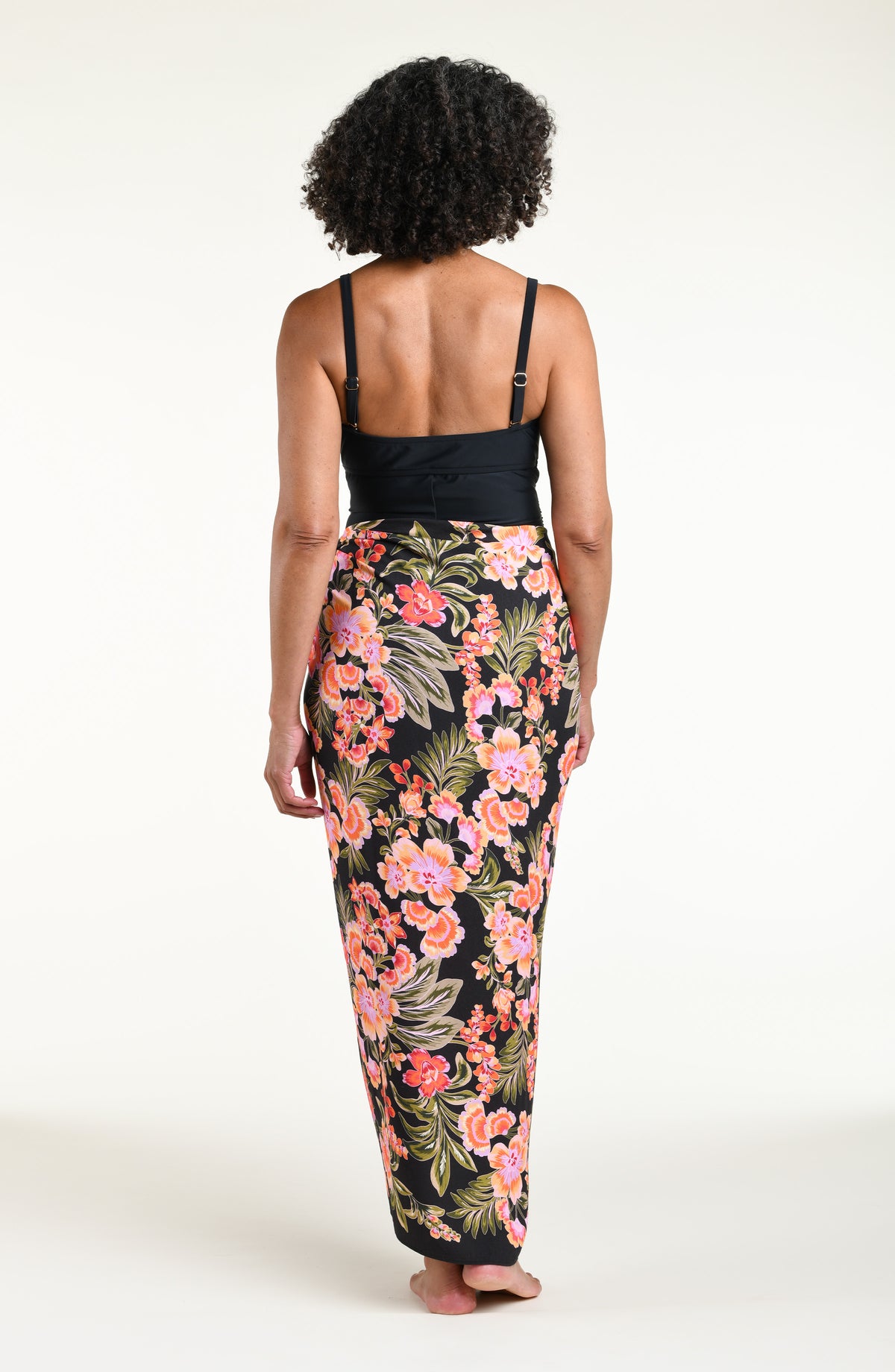 A woman modeling a black one piece swimsuit and a wrap sarong with a vibrant pink, orange, and green floral print, featuring a high side slit and a tied waist.