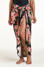 A woman modeling a black wrap sarong with a vibrant pink, orange, and green floral print, featuring a high side slit and a tied waist.