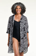 A woman modeling a solid black one piece swimsuit under a black and white tropical leaf print tunic with three-quarter sleeves and a high-low hem, shown against a plain white studio background.