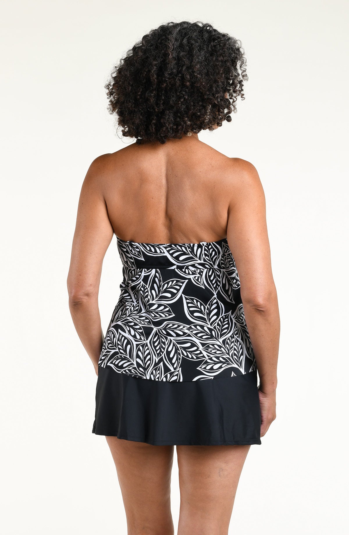 A woman wearing a black and white tropical leaf print camisole top and a black swim skirt, standing against a plain off-white studio background.