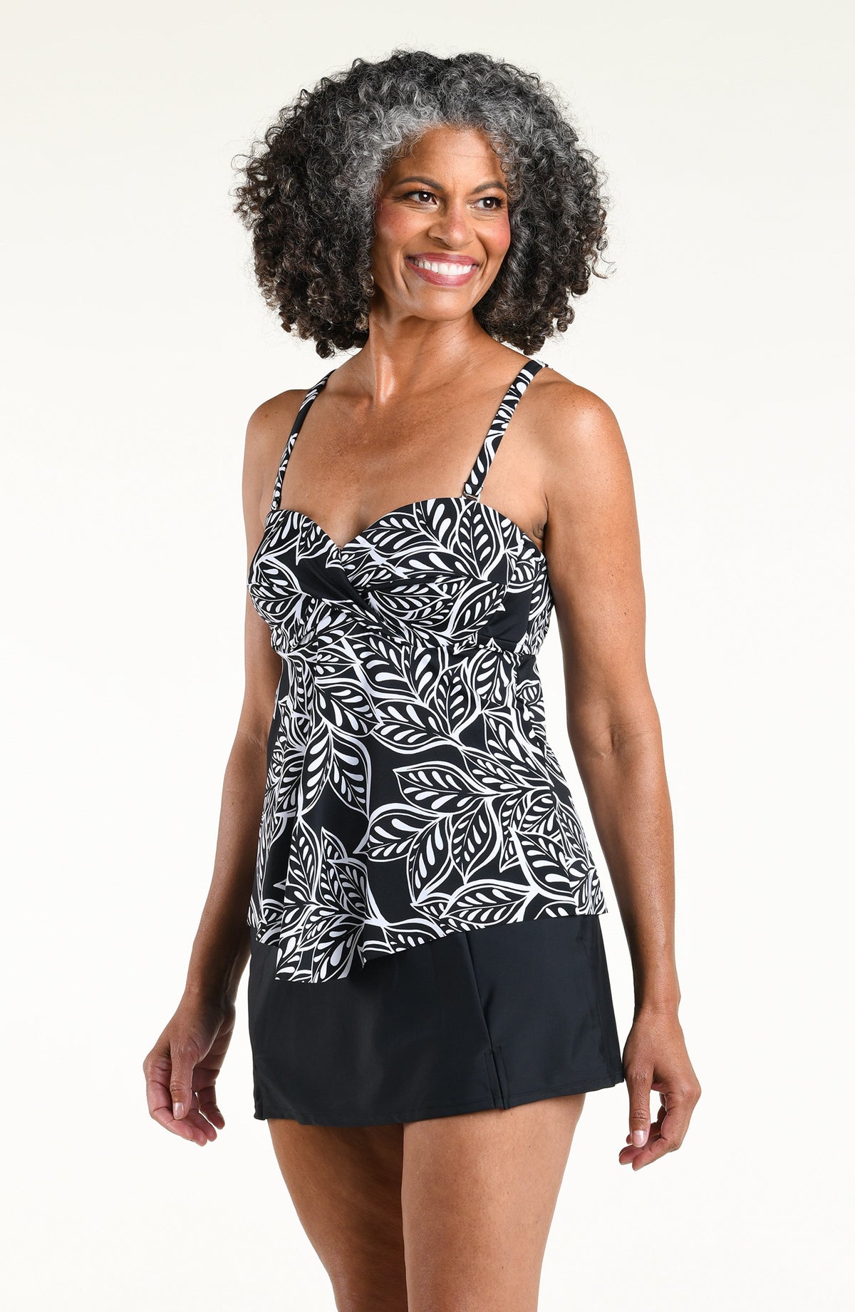 A woman wearing a black and white tropical leaf print camisole top and a black swim skirt, standing against a plain off-white studio background.