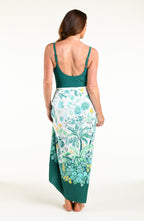 A woman modeling a two-piece swimwear set consisting of a solid teal green tankini top and a matching teal green tropical floral print sarong skirt, set against a plain white studio background.