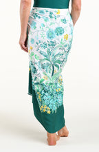 A woman models a high-low wrap sarong with a green and white botanical print, featuring tassel ties and a side slit, shown against a plain light background.