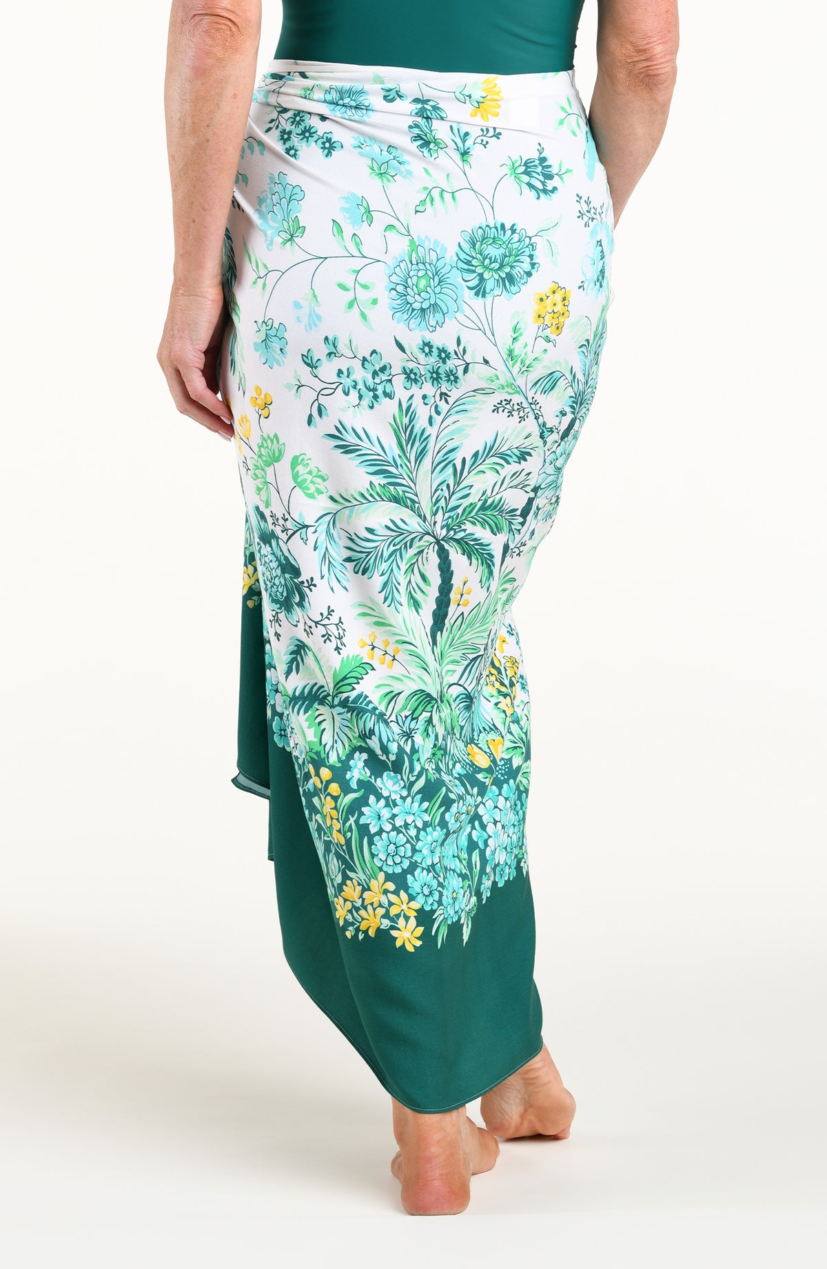 A woman models a high-low wrap sarong with a green and white botanical print, featuring tassel ties and a side slit, shown against a plain light background.