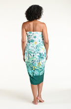 A woman models a high-low wrap sarong with a green and white botanical print, featuring tassel ties and a side slit, shown against a plain light background.