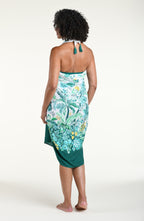 A woman models a high-low wrap sarong with a green and white botanical print, featuring tassel ties and a side slit, shown against a plain light background.