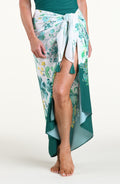 A woman models a high-low wrap sarong with a green and white botanical print, featuring tassel ties and a side slit, shown against a plain light background.