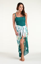 A woman modeling a two-piece swimwear set consisting of a solid teal green tankini top and a matching teal green tropical floral print sarong skirt, set against a plain white studio background.