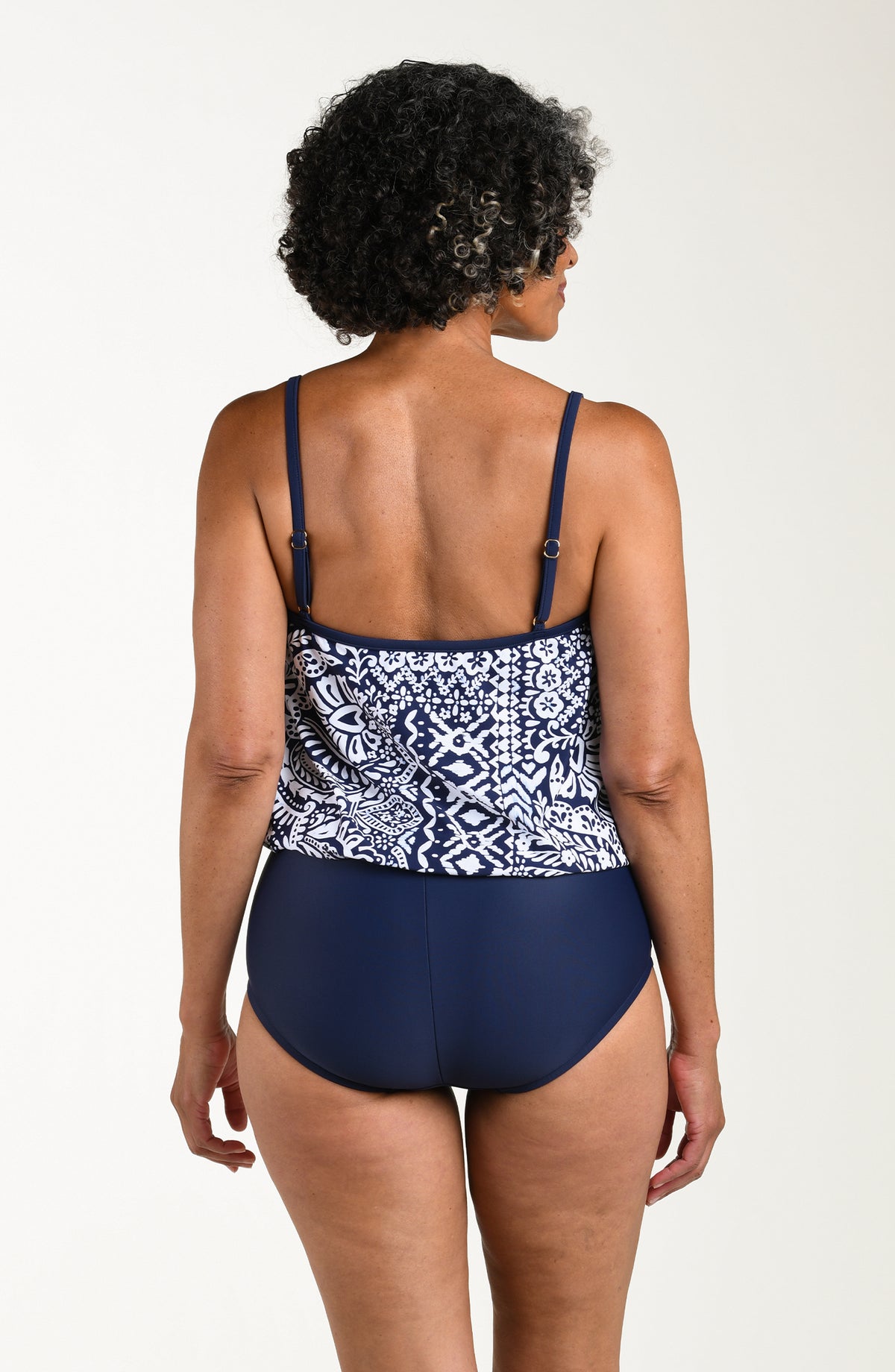 Back image of model wearing a multicolored navy blue paisley printed one-piece swimsuit featuring thin straps, a straight back, and a loose-flowing overlay. The top looks like a tankini and the bottom is solid indigo (navy blue) colored.