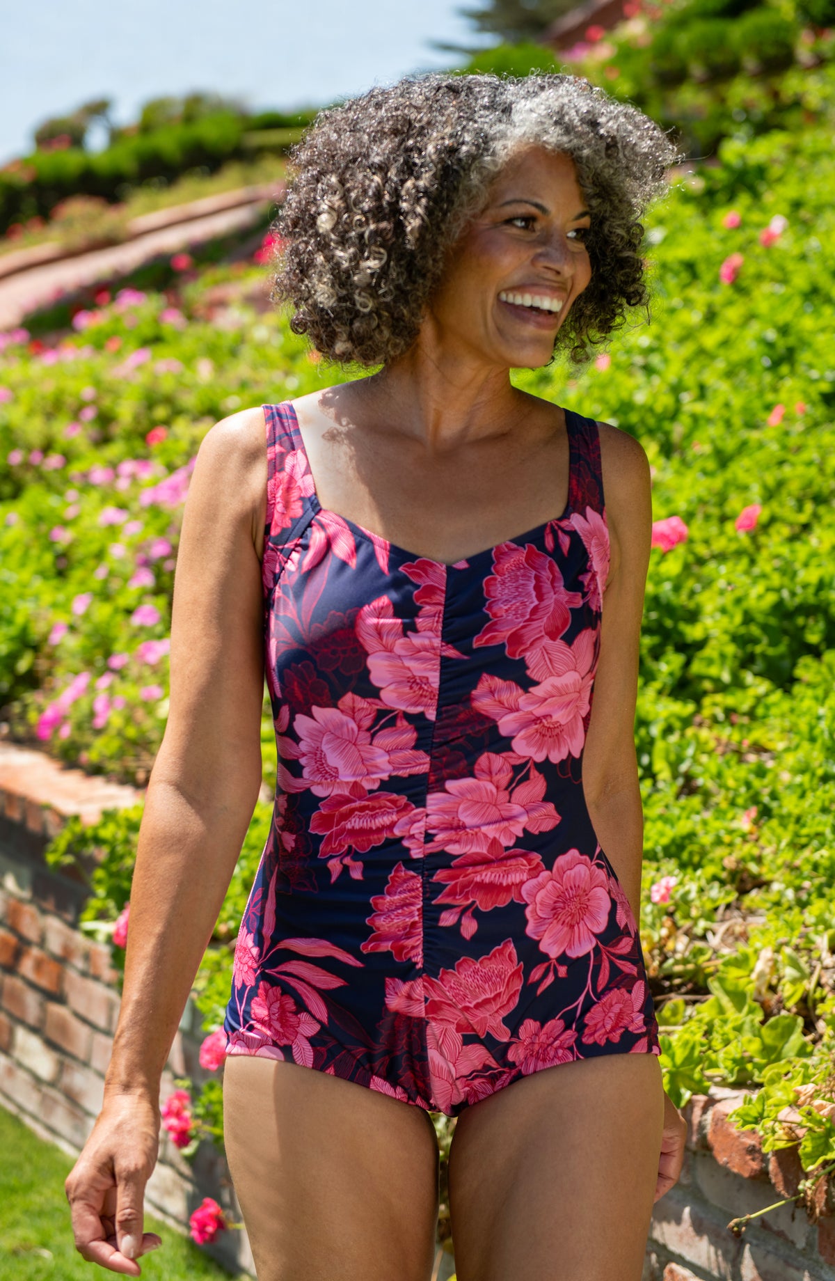 Lifestyle front image of model wearing a multicolored (pink) floral printed one-piece swimsuit with wide straps, a sweetheart neckline, ruched body, and girl let cut. 