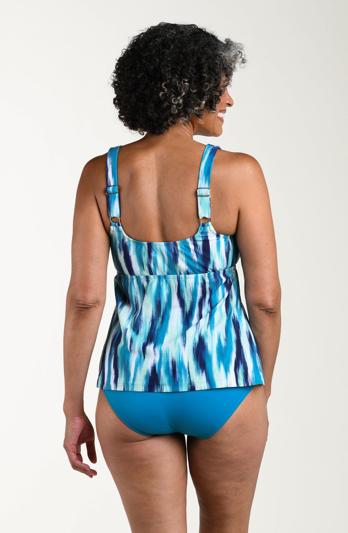 Back image of model wearing a multicolored teal tankini featuring a scoop neckline, thick over the shoulder straps, and empire waist.