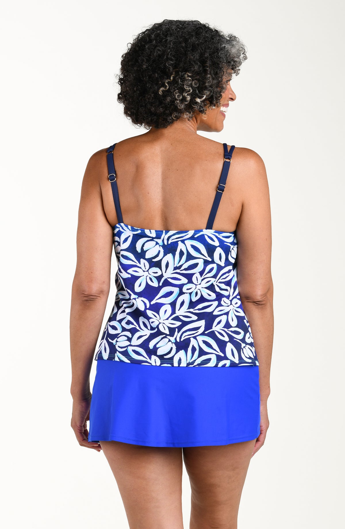 Back image of model wearing a multicolored floral printed tankini top featuring a adjustable shoulder straps, and a straight back. This is paired with a cobalt colored swim skirt. 