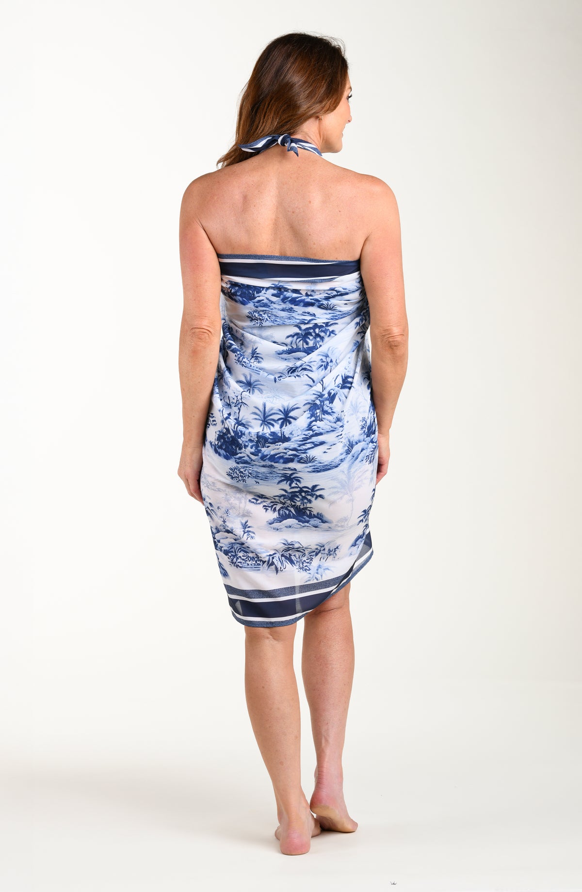 Full body back image of model wearing a multicolored white and blue tropical pareo which is tied at the neckline. 