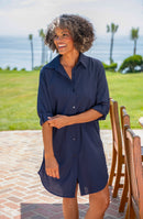 Lifestyle front image of model wearing a solid indigo colored button-down swimsuit cover-up with a collared neckline, rolled 3/4 sleeves, and a mid-thigh length.